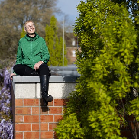 Frau sitzt umgeben von grünen Pflanzen lachend auf Mauer.