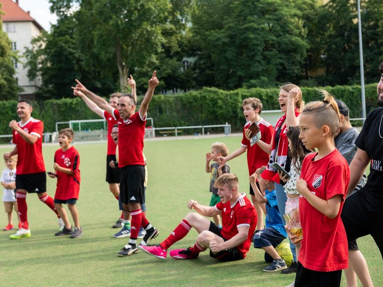 Kinder, Jugendliche und Männer in Trikots jubeln am Spielfeldrand. 