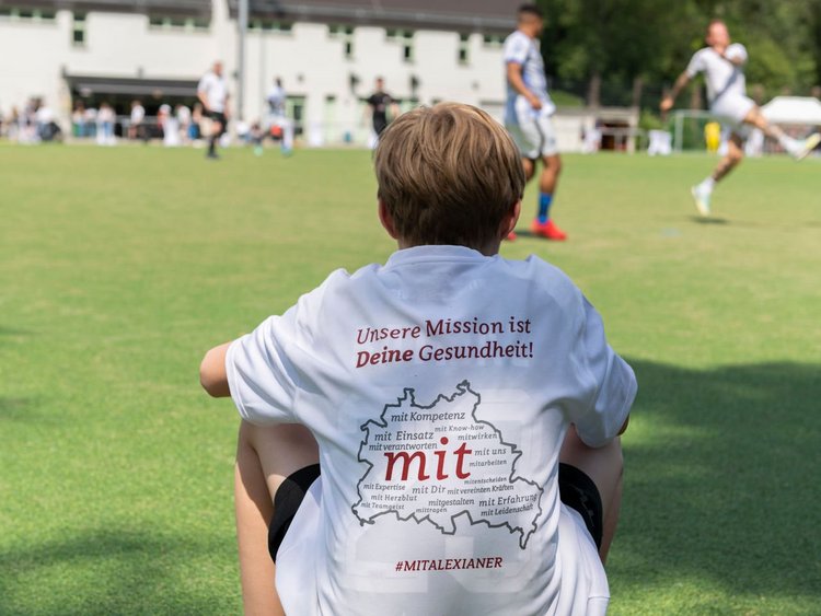 Junge sitzt auf einem Fußball am Spielfeldrand und beobachtet das Spiel